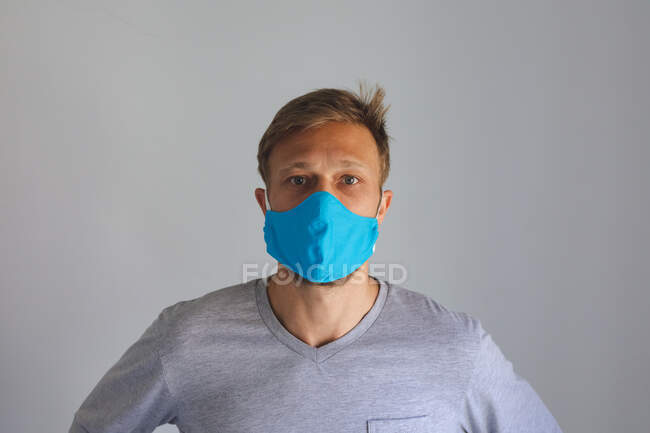 Ritratto di uomo caucasico che trascorre del tempo a casa, indossando una maschera blu che guarda la macchina fotografica su sfondo grigio. Distanza sociale durante il blocco di quarantena Covid 19 Coronavirus. — Foto stock