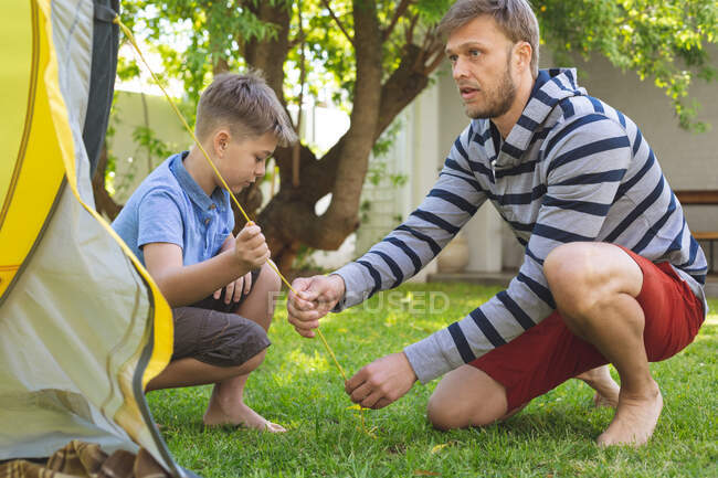 Hombre caucásico pasando tiempo con su hijo juntos, acampando en el jardín, poniendo tienda de campaña. Distanciamiento social durante el bloqueo de cuarentena del Coronavirus Covid 19. - foto de stock