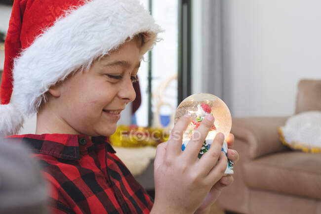 Белый мальчик дома на Рождество, сидит на диване в гостиной в шляпе Санты, держа снежный шар. Социальное дистанцирование во время изоляции коронавируса Covid 19. — стоковое фото