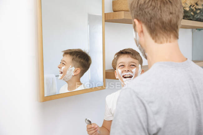 Caucasian man at home with his son together, in bathroom, shaving with shaving cream on faces, smiling. Social distancing during Covid 19 Coronavirus quarantine lockdown. — Stock Photo