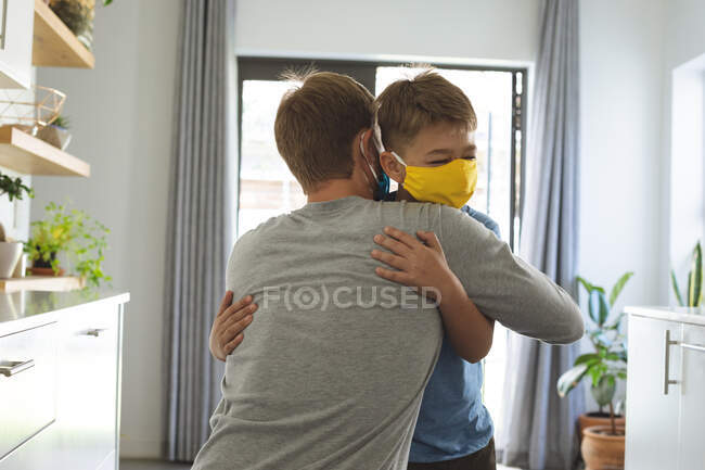Hombre caucásico en casa con su hijo, en la cocina, con mascarillas, abrazos. Distanciamiento social durante el bloqueo de cuarentena del Coronavirus Covid 19. - foto de stock