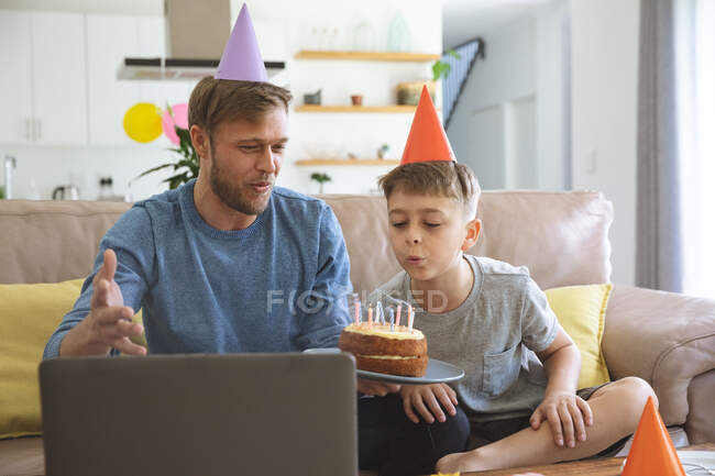 Hombre caucásico pasar tiempo en casa con su hijo juntos, soplando velas en la torta de cumpleaños utilizando el ordenador portátil para el chat de vídeo. Distanciamiento social durante el bloqueo de cuarentena del Coronavirus Covid 19. - foto de stock