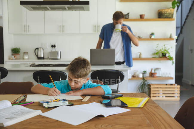 Uomo caucasico a casa con suo figlio insieme, in cucina, ragazzo che fa i compiti a tavola. Distanza sociale durante il blocco di quarantena Covid 19 Coronavirus. — Foto stock