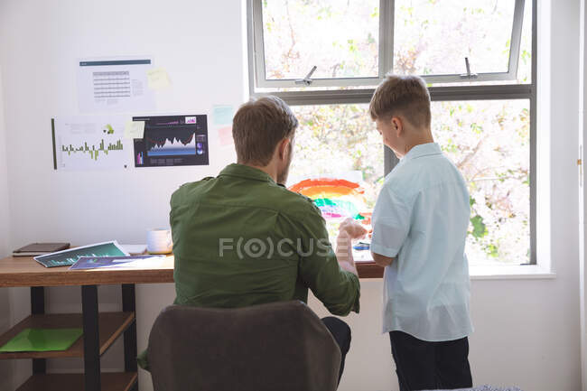 Белый мужчина дома со своим сыном вместе, за столом, помогая с домашним обучением. Социальное дистанцирование во время изоляции коронавируса Covid 19. — стоковое фото