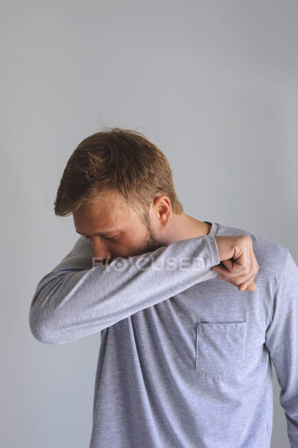 Caucasian man spending time at home, being sick, coughing sneezing, covering his face with elbow on grey background. Social distancing during Covid 19 Coronavirus quarantine lockdown. — Stock Photo