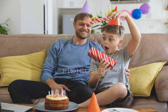 Caucasian man spending time at home with his son together, wearing party hats boy receiving birthday present. Social distancing during Covid 19 Coronavirus quarantine lockdown. — Stock Photo