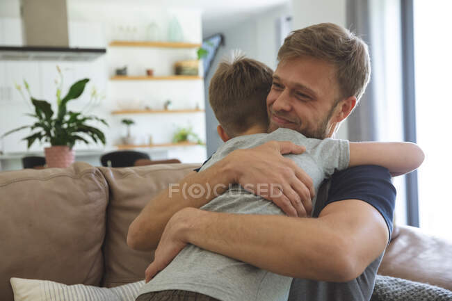 Hombre caucásico en casa con su hijo juntos, sentado en el sofá en la sala de estar, abrazándose, sonriendo. Distanciamiento social durante el bloqueo de cuarentena del Coronavirus Covid 19. - foto de stock