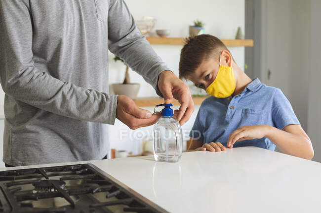 Homme caucasien à la maison avec son fils, dans la cuisine, garçon portant un masque facial, homme désinfectant ses mains. Distance sociale pendant le confinement en quarantaine du coronavirus Covid 19. — Photo de stock