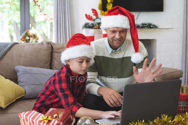 Caucasian man at home with his son at Christmas, wearing Santa hats sitting in living room using laptop for video call. Social distancing during Covid 19 Coronavirus quarantine lockdown. — Stock Photo