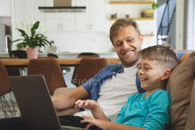 Hombre caucásico en casa con su hijo juntos, sentado en el sofá en la sala de estar, utilizando el ordenador portátil, sonriendo. Distanciamiento social durante el bloqueo de cuarentena del Coronavirus Covid 19. - foto de stock