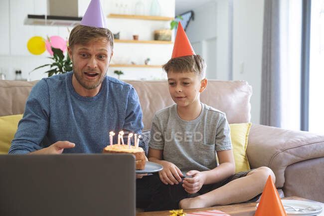 Hombre caucásico pasar tiempo en casa con su hijo juntos, teniendo fiesta de cumpleaños utilizando el ordenador portátil para el chat de vídeo. Distanciamiento social durante el bloqueo de cuarentena del Coronavirus Covid 19. - foto de stock