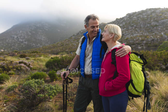 Coppia anziana trascorrere del tempo in natura insieme, passeggiando in montagna, l'uomo sta abbracciando la donna, guardando e l'un l'altro e sorridendo. stile di vita sano attività pensionistica. — Foto stock