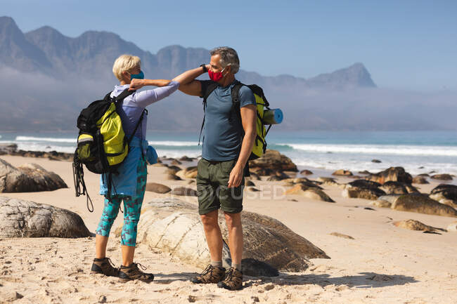 Coppia anziana trascorrere del tempo in natura insieme, passeggiare sulla spiaggia, indossare maschere per il viso e salutarsi con i gomiti. stile di vita sano attività pensionistica. — Foto stock