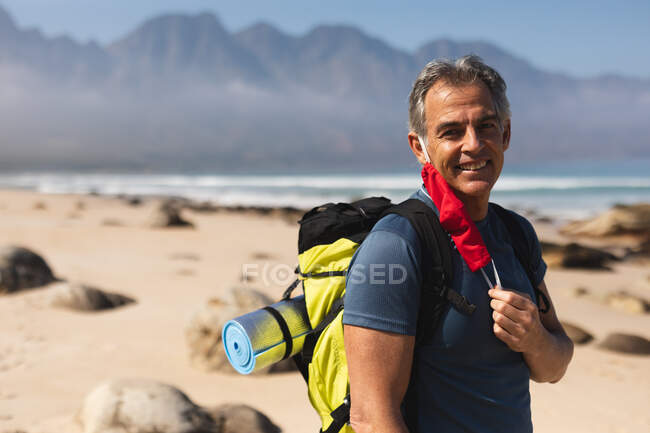 Porträt eines älteren Mannes, der Zeit in der Natur verbringt, am Strand spazieren geht, eine Gesichtsmaske aufsetzt, in die Kamera schaut und lächelt. Gesunder Lebensstil im Ruhestand. — Stockfoto