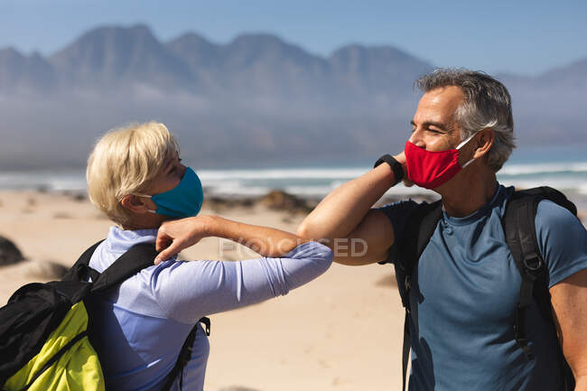 Casal sênior passando tempo na natureza juntos, andando na praia, vestindo máscaras e cumprimentando uns aos outros com cotovelos. atividade de aposentadoria saudável. — Fotografia de Stock