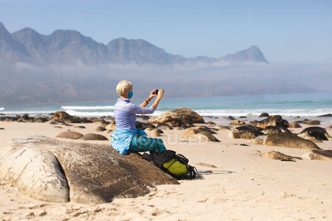 Старшая женщина проводит время на природе, гуляет по пляжу, носит маску для лица и делает селфи. активный уход на пенсию. — стоковое фото