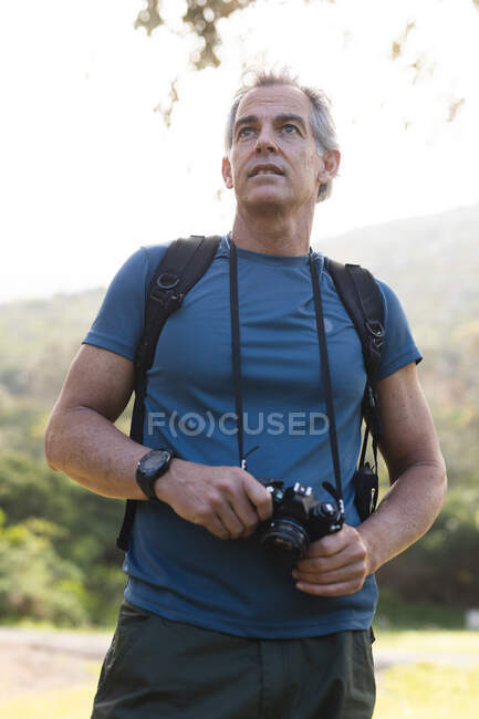 Hombre mayor pasando tiempo en la naturaleza, caminando por las montañas, sosteniendo una cámara. estilo de vida saludable actividad de jubilación. - foto de stock