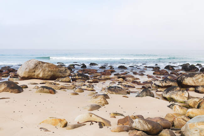 Magnífica vista de um mar calmo e uma praia com areia amarela e rochas bastante grandes deitadas na praia. — Fotografia de Stock