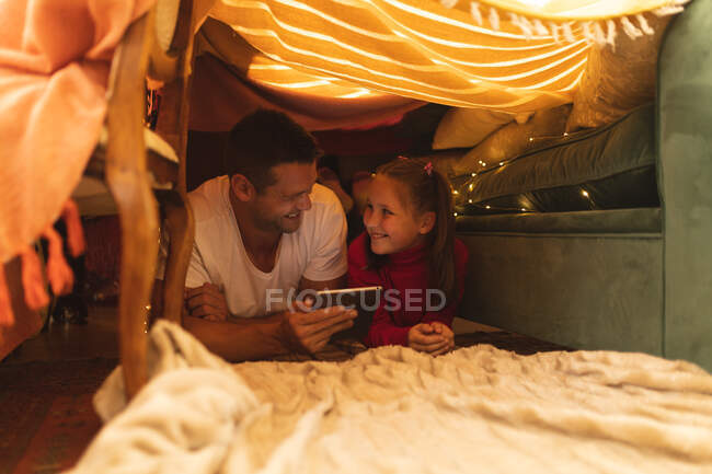 Кавказский мужчина с дочерью улыбается и пользуется табличкой, лежа в одеяле форта. качественный образ жизни. — стоковое фото
