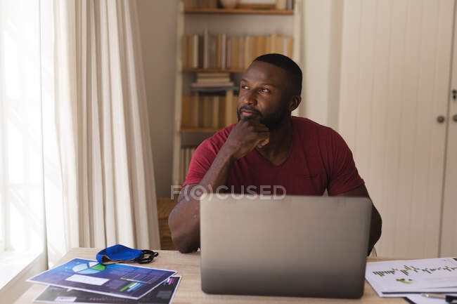 Thoughtful  african american man looking out of window with laptop, face mask and documents on table at home. social distancing during covid 19 coronavirus quarantine lockdown. — Stock Photo