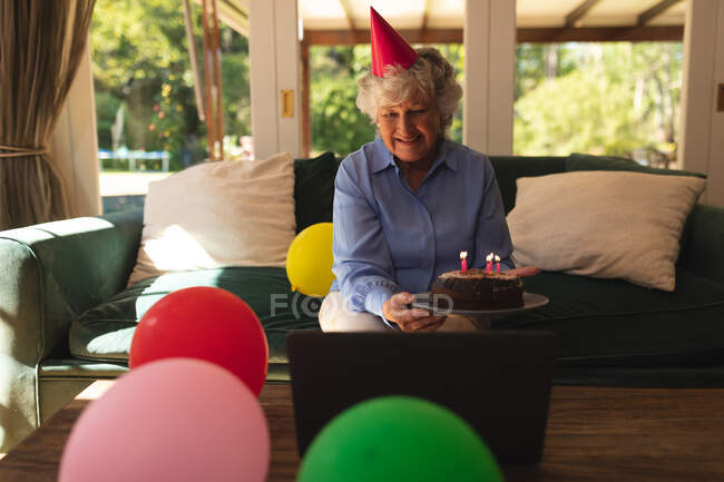 Eine ältere kaukasische Frau, die zu Hause einen Geburtstag feiert, einen Partyhut trägt und einen Laptop benutzt. Selbstisolation zu Hause während Coronavirus covid 19 Quarantäne Lockdown. — Stockfoto