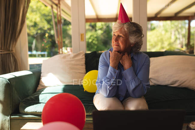 Старша жінка-кавказ проводить час удома, відзначаючи день народження, одягаючи капелюх на вечірці і використовуючи ноутбук. Самоізоляція вдома під час коронавірусу коїда 19 карантин.. — стокове фото