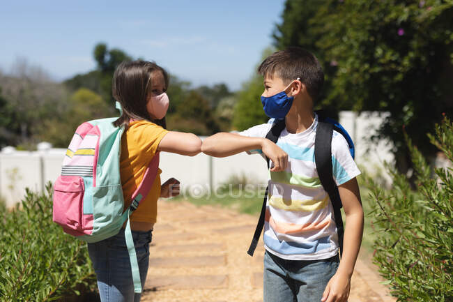 Кавказький хлопчик і дівчинка в масках на обличчі вітаються по дорозі до школи, торкаючись ліктів. освіта і спосіб життя під час пандемії коронавірусу 19 — стокове фото