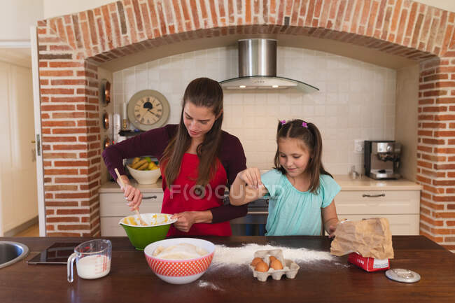 Mujer caucásica con su hija horneando en una cocina y usando delantales. auto aislamiento en casa durante coronavirus covid 19 bloqueo de cuarentena. - foto de stock