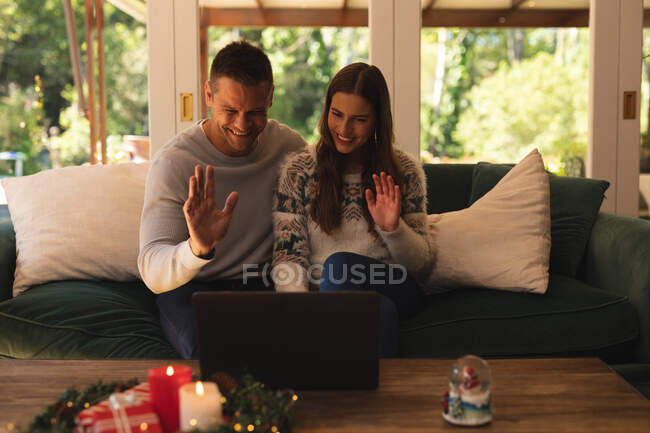 Casal caucasiano acenando as mãos e sorrindo, fazendo uma chamada de vídeo usando laptop na época do Natal. auto-isolamento durante o bloqueio de quarentena do coronavírus covid 19. — Fotografia de Stock