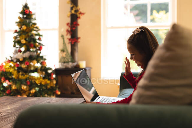 Menina caucasiana sentada no sofá, acenando e sorrindo, fazendo uma chamada de vídeo usando um laptop na época do Natal. auto-isolamento durante o bloqueio de quarentena do coronavírus covid 19. — Fotografia de Stock