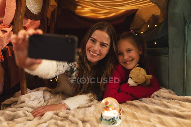 Kaukasische Frau und ihre Tochter lächeln und machen Selfie mit Smartphone, in einer Decke liegend, das Mädchen umarmt Teddybär. Selbstisolation während Coronavirus covid 19 Quarantäne Lockdown. — Stockfoto