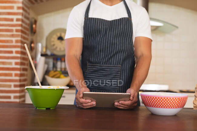 Hombre de pie en una cocina y usando delantal, usando su tableta. auto aislamiento en casa durante coronavirus covid 19 bloqueo de cuarentena. - foto de stock