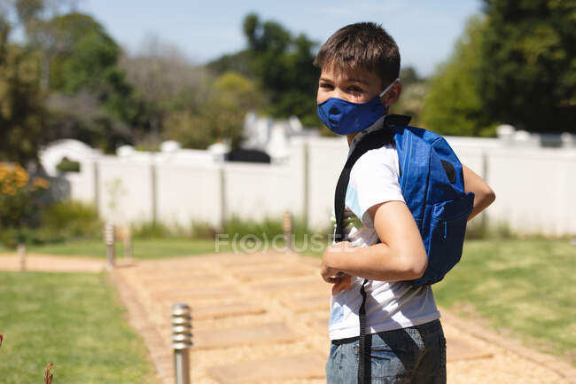 Retrato de niño caucásico con máscara facial mirando a la cámara de pie en el jardín con la mochila en el día soleado. protección y autoaislamiento durante el bloqueo pandémico del coronavirus covid 19. - foto de stock