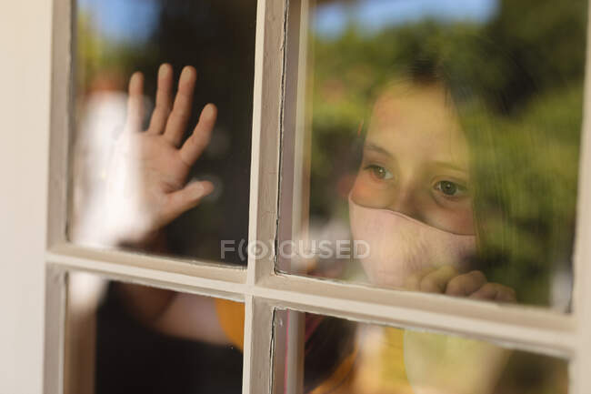 Menina caucasiana usando máscara facial e olhando através da janela. auto-isolamento durante o bloqueio de quarentena do coronavírus covid 19. — Fotografia de Stock