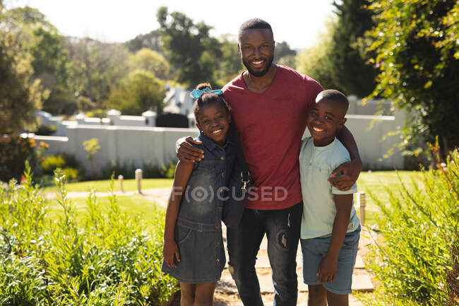 Retrato de padre, hija e hijo afroamericanos sonriendo mientras estaban en el jardín en un día soleado brillante. distanciamiento social durante el bloqueo de cuarentena por coronavirus covid 19 - foto de stock