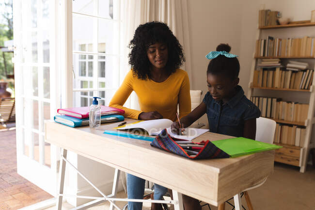 Mãe afro-americana ajudando filha com lição de casa enquanto sentada em cadeiras em casa. distanciamento social durante o bloqueio de quarentena do coronavírus covid 19. — Fotografia de Stock