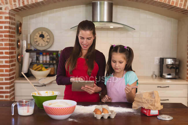 Белая женщина с дочерью пекут на кухне и носят фартуки, используя планшет. самоизоляция дома во время блокады коронавируса 19. — стоковое фото