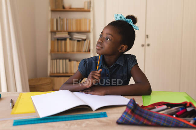 Chica afroamericana sosteniendo lápiz mirando por la ventana con libros, pluma y regla en la mesa en casa. distanciamiento social durante el bloqueo de cuarentena por coronavirus covid 19. - foto de stock