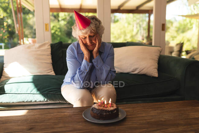 Eine ältere kaukasische Frau, die zu Hause einen Geburtstag feiert, einen Partyhut trägt und sich Kuchen ansieht. Selbstisolation zu Hause während Coronavirus covid 19 Quarantäne Lockdown. — Stockfoto