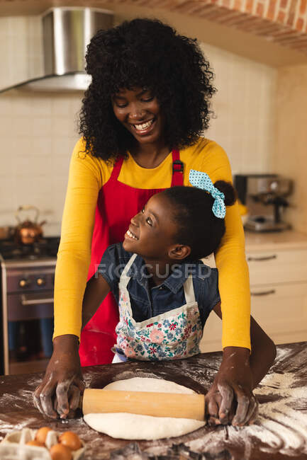 Madre e hija afroamericanas vistiendo delantales sonriendo mientras se miran en la cocina en casa. fiesta de navidad tradición celebración concepto - foto de stock