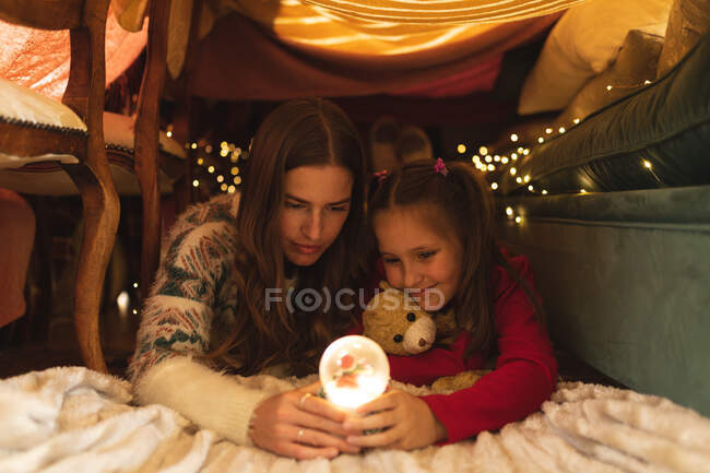 Mulher caucasiana e sua filha sorrindo e segurando globo de neve, deitada no forte cobertor. auto-isolamento durante o bloqueio de quarentena do coronavírus covid 19. — Fotografia de Stock