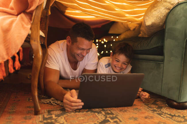 Hombre caucásico con su hijo sonriendo y usando tableta, acostado en un fuerte de mantas. estilo de vida de calidad tiempo familiar juntos. - foto de stock