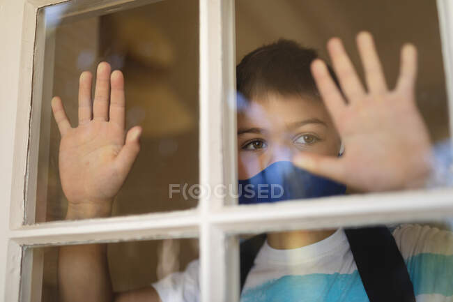 Caucasian boy looking through window and wearing face mask. self isolation at home during coronavirus covid 19 quarantine lockdown. — Stock Photo
