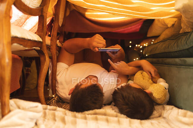 Kaukasischer Mann mit seinem Sohn, der Tabletten nimmt und in einer Deckenfestung liegt. qualitativ hochwertiger Lebensstil Familienzeit zusammen. — Stockfoto