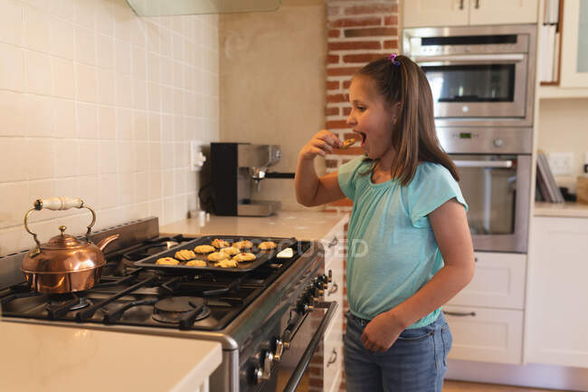 Chica caucásica de pie en una cocina y comer galletas de bandeja para hornear. auto aislamiento en casa durante coronavirus covid 19 bloqueo de cuarentena. - foto de stock
