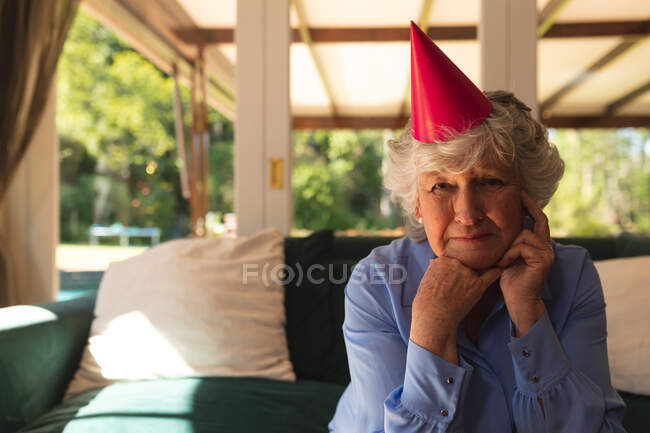 Портрет старшої кавказької жінки проводить час удома, відзначаючи день народження, одягаючи капелюх на вечірку і дивлячись на камеру. Самоізоляція вдома під час коронавірусу коїда 19 карантин.. — стокове фото