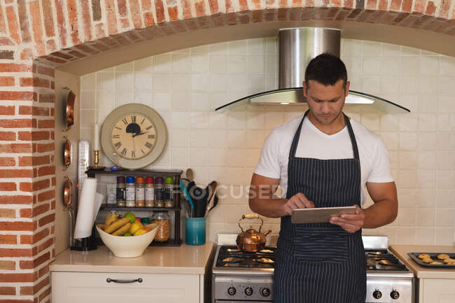 Белый мужчина стоит на кухне в фартуке и пользуется планшетом. самоизоляция во время блокады коронавируса 19. — стоковое фото