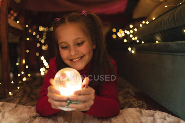 Chica caucásica sonriendo y sosteniendo bola de nieve, acostada en el fuerte manta. autoaislamiento durante el bloqueo de cuarentena del covid 19 del coronavirus. - foto de stock