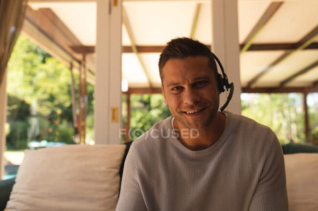 Retrato de un hombre caucásico trabajando desde casa con un auricular de teléfono sentado en la sala de estar y sonriendo a la cámara. autoaislamiento durante la pandemia de coronavirus covid 19. - foto de stock