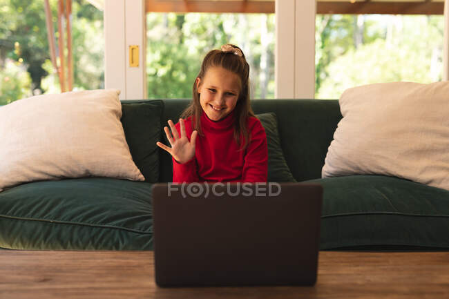 Белая девушка сидит на диване, машет и улыбается, делает видеозвонок с помощью ноутбука. самоизоляция во время блокады коронавируса 19. — стоковое фото